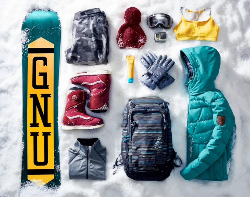 Snowboard, jacket, and cold weather gear arranged on snow. Flat lay of snowboard, backpack, jacket, goggles, gloves, and other winter apparel.