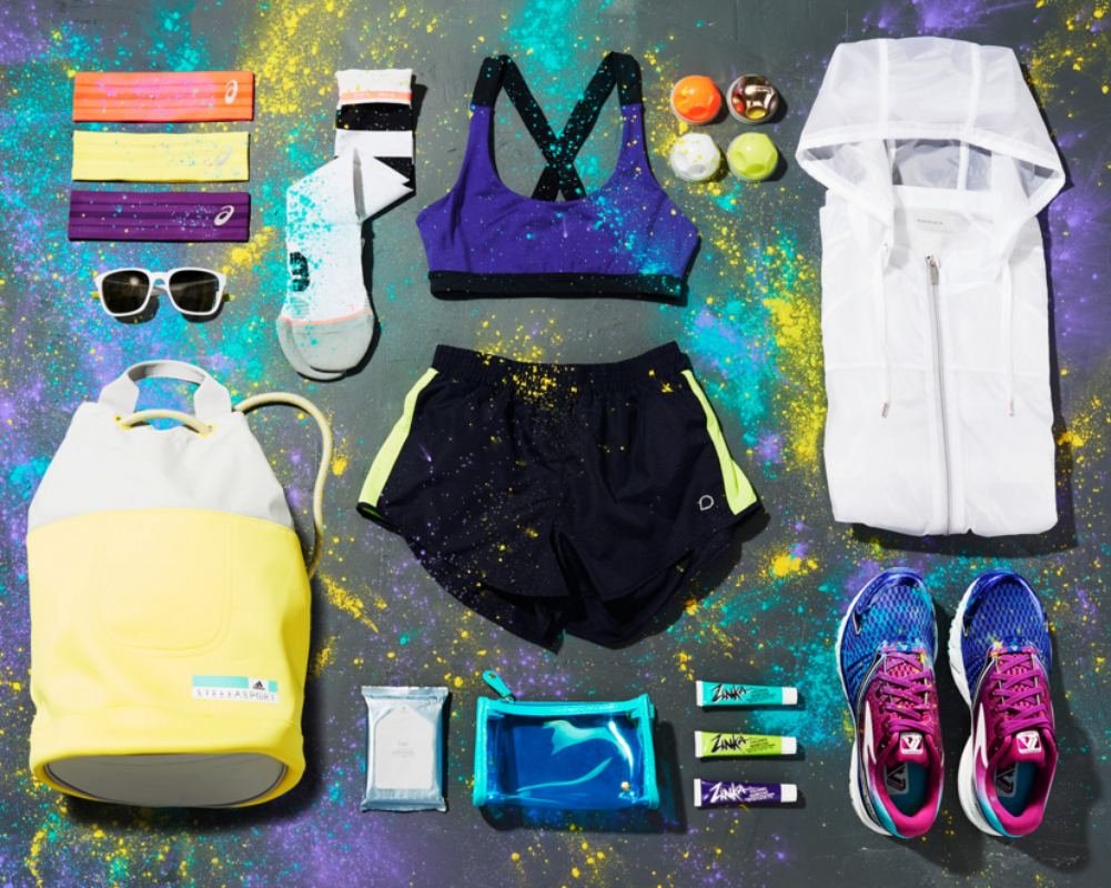 Flat lay of sports gear including running shoes, jacket, shorts, sports bra, socks, sunglasses, and accessories laid out on a gray surface.