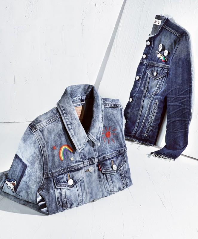 Two denim jackets, one with a rainbow and sun design, are on a white surface, one hanging, the other folded, lit by bright studio light.