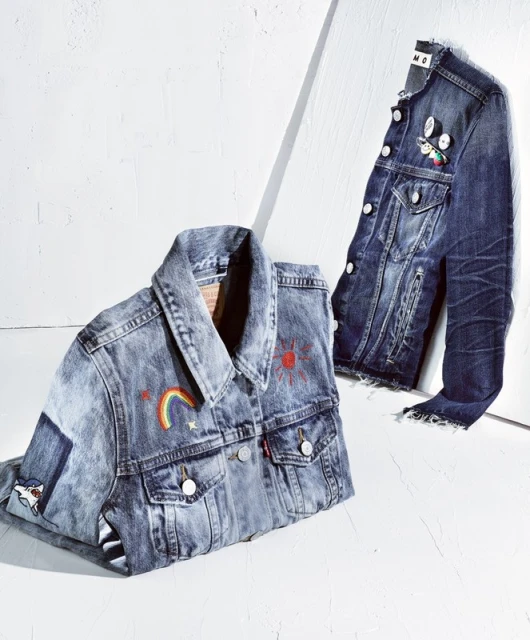 Two denim jackets, one with a rainbow and sun design, are on a white surface, one hanging, the other folded, lit by bright studio light.