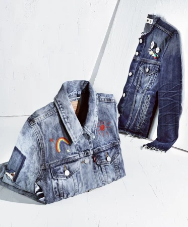 Two denim jackets, one with a rainbow and sun design, are on a white surface, one hanging, the other folded, lit by bright studio light.