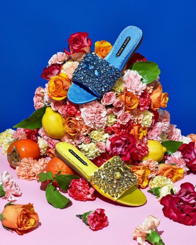 Two embellished Alice + Olivia sandals on a bed of roses and assorted fruit, set against a blue and pink backdrop in a studio setting.