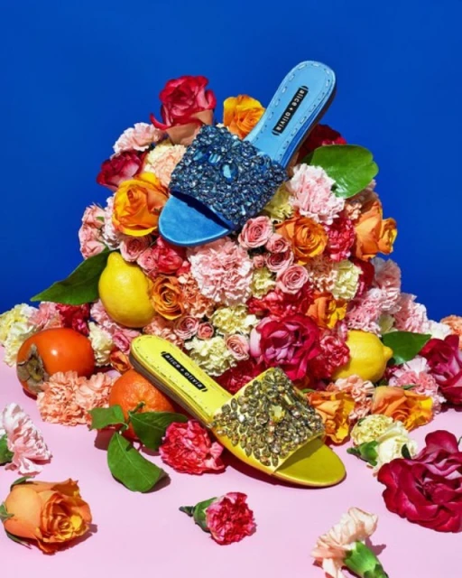 Two embellished Alice + Olivia sandals on a bed of roses and assorted fruit, set against a blue and pink backdrop in a studio setting.
