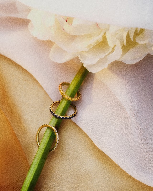 Close-up of three gold rings stacked on the stem of a white flower, set against a soft, draped fabric backdrop. Jewellery advertisement.