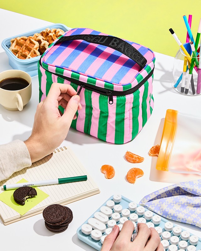 Top-down shot of a striped BAGGUGU lunchbox, coffee cup, keyboard, and person's hands on a white desk with yellow background.
