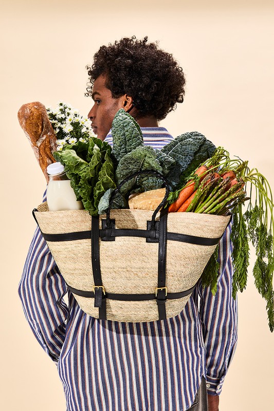 Person with curly hair and striped shirt carries a straw tote filled with kale, asparagus, carrots, baguette, and flowers in studio.