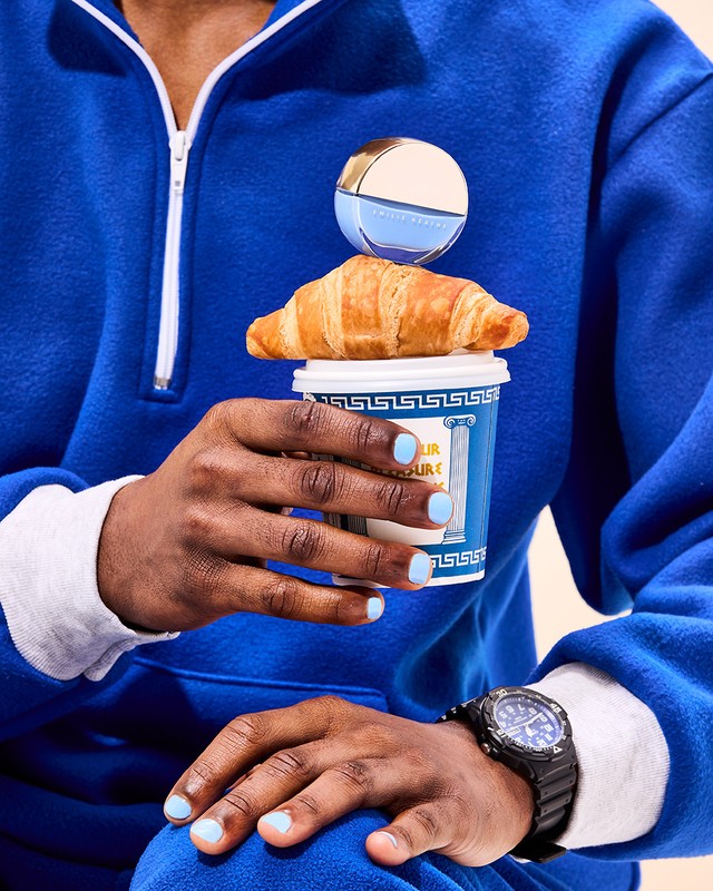 Close-up shot of an individual in a blue sweatshirt, holding a coffee cup topped with a croissant and perfume bottle, and wearing a watch.