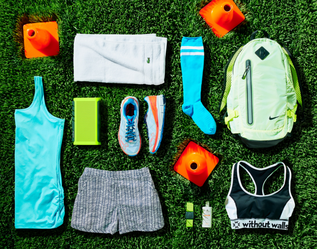 Overhead shot of neatly arranged fitness gear on green grass: running shoes, turquoise top, sports bra, backpack, and cones.