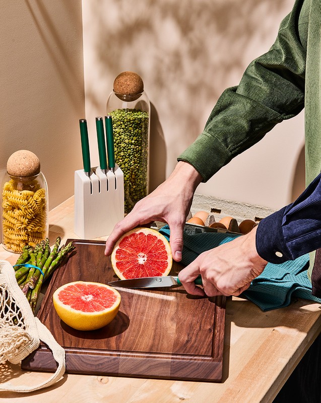 A person slices a grapefruit on a wooden cutting board in a kitchen setting, alongside ingredients like pasta, lentils, and asparagus.