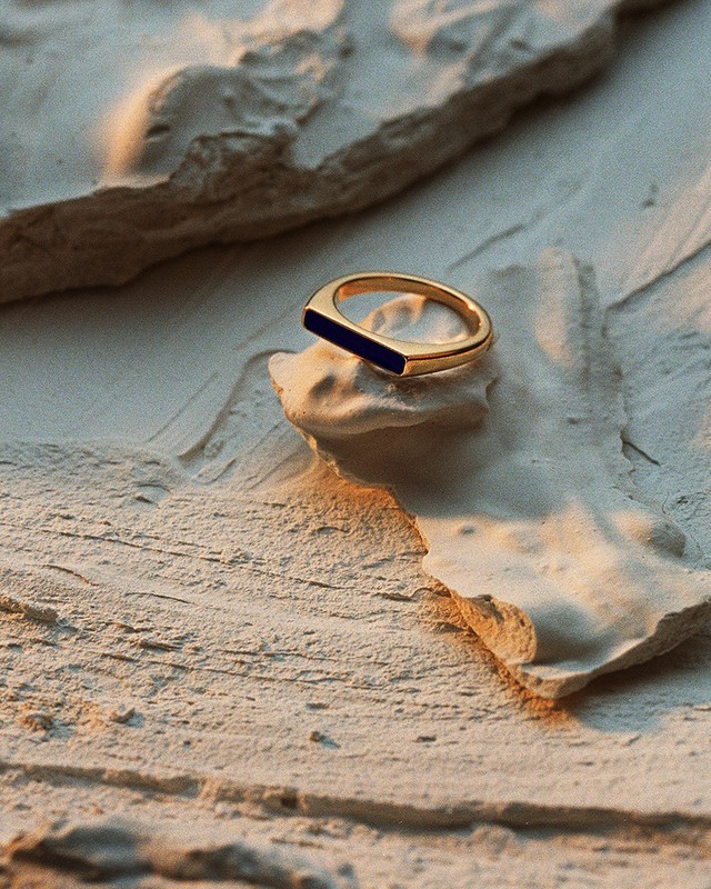 Close-up of a gold ring with a blue stone on a textured, light-colored stone surface. Jewelry photography.