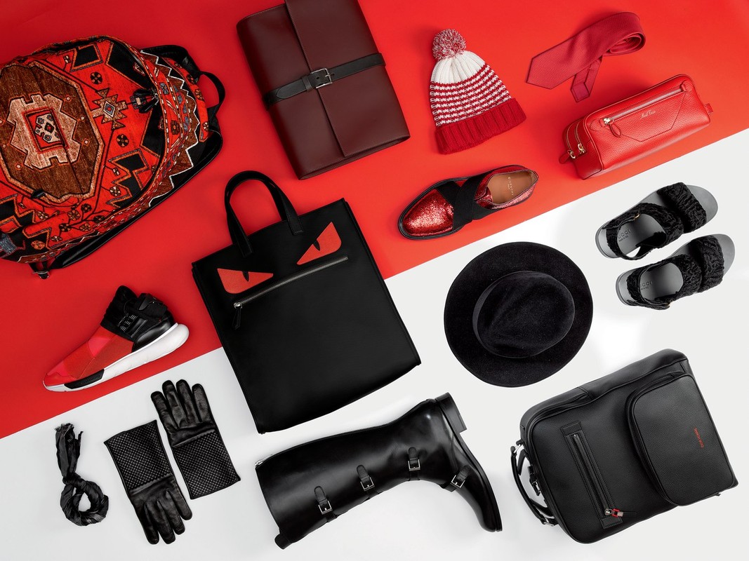 Flat lay of fashion and travel accessories, including a backpack, tote bag, shoes, boots, hat, tie, gloves, and other items on a red and white background.