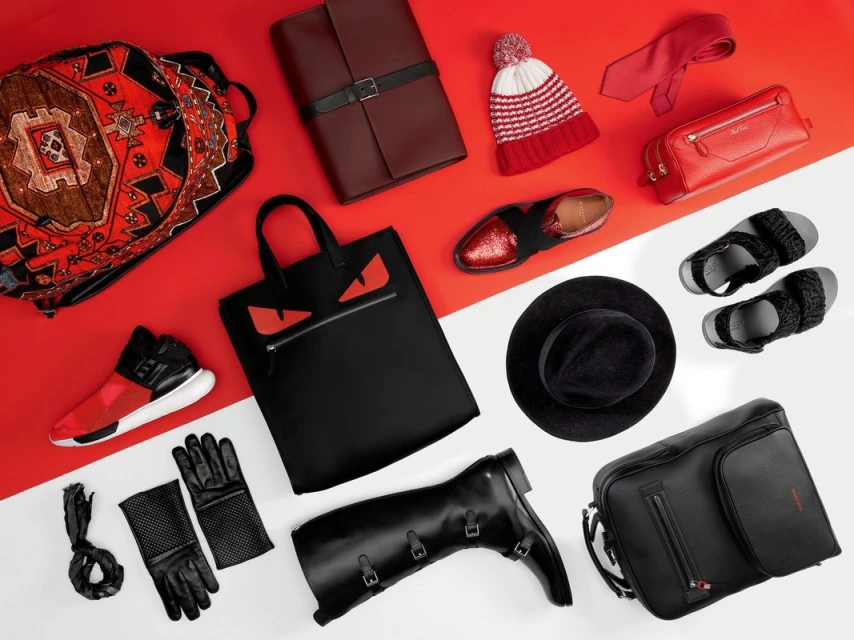 Flat lay of fashion and travel accessories, including a backpack, tote bag, shoes, boots, hat, tie, gloves, and other items on a red and white background.
