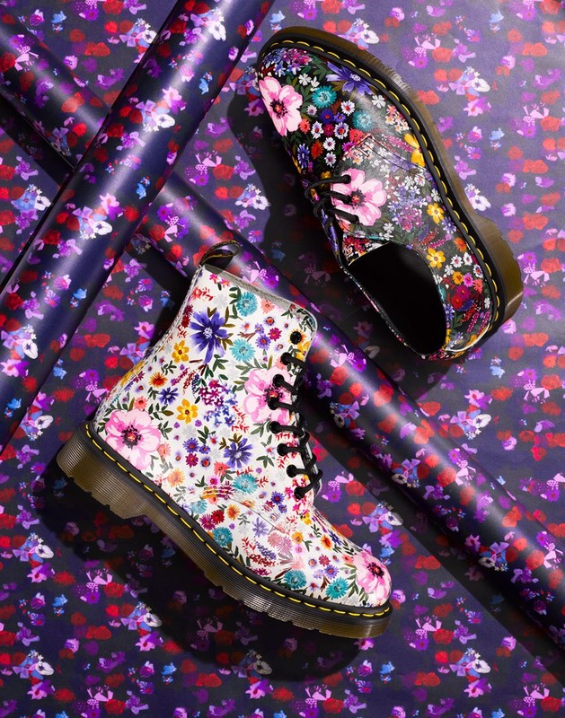 Floral-print Dr. Martens boots on a matching floral background with parallel purple tubes diagonally across the frame.