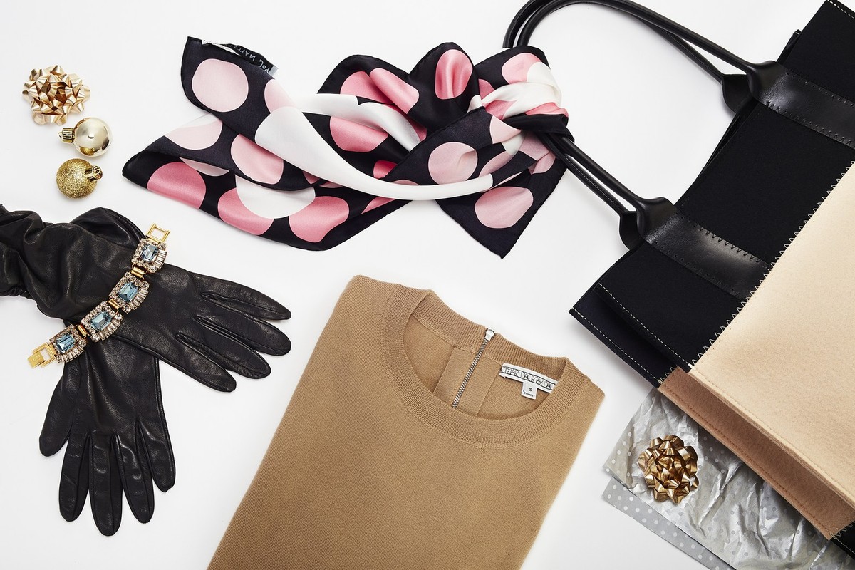 Flat lay of a beige sweater, black leather gloves with a jeweled bracelet, and a tote bag against a white backdrop.