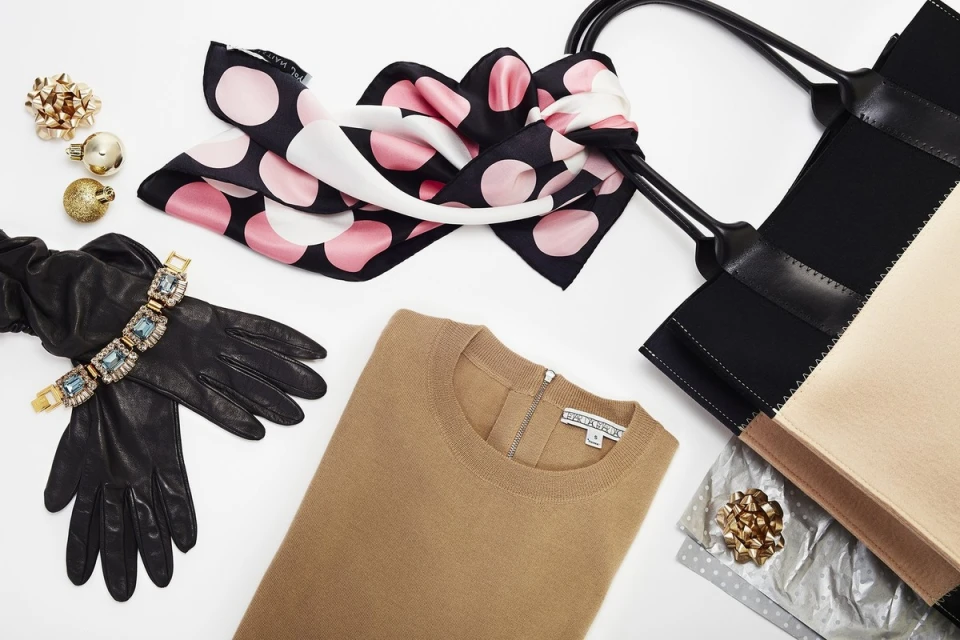 Flat lay of a beige sweater, black leather gloves with a jeweled bracelet, and a tote bag against a white backdrop.