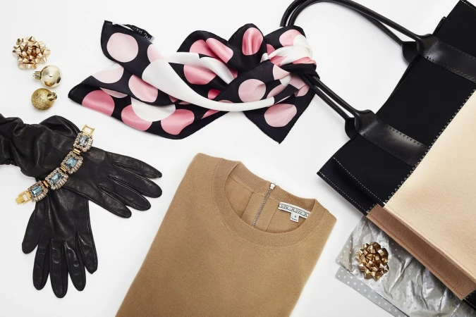 Flat lay of a beige sweater, black leather gloves with a jeweled bracelet, and a tote bag against a white backdrop.