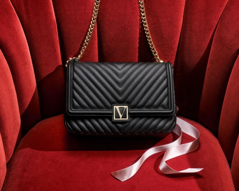 Elegant black shoulder bag with gold chain on red velvet chair, featuring a light pink ribbon accent for luxury fashion appeal.