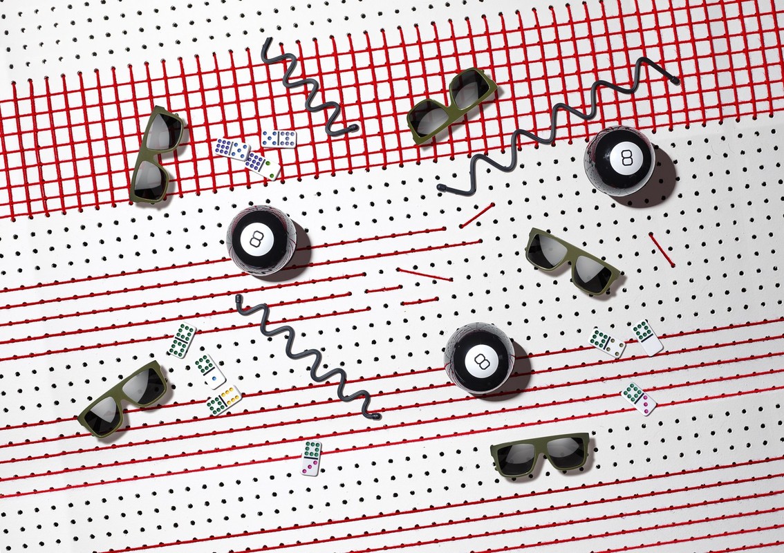 Arrangement of sunglasses, billiard balls, and dominoes on a white surface with red lines and black dots in a studio setting.