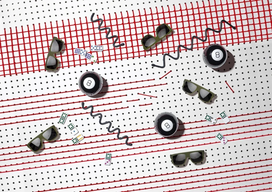 Arrangement of sunglasses, billiard balls, and dominoes on a white surface with red lines and black dots in a studio setting.