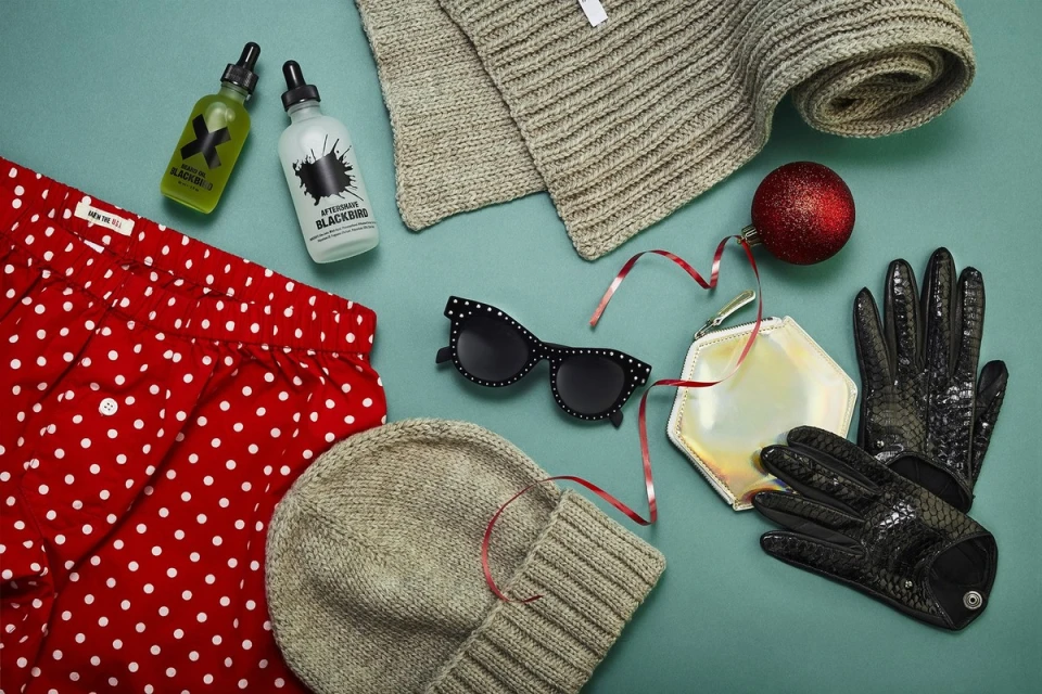 Flat lay of various winter accessories and beauty products on a blue surface, including a scarf, hat, gloves, and polka dot patterned shorts.