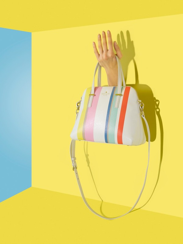 Stylish handbag with colorful stripes held by a hand against a yellow wall with blue corner, creating a vibrant and eye-catching fashion still life.