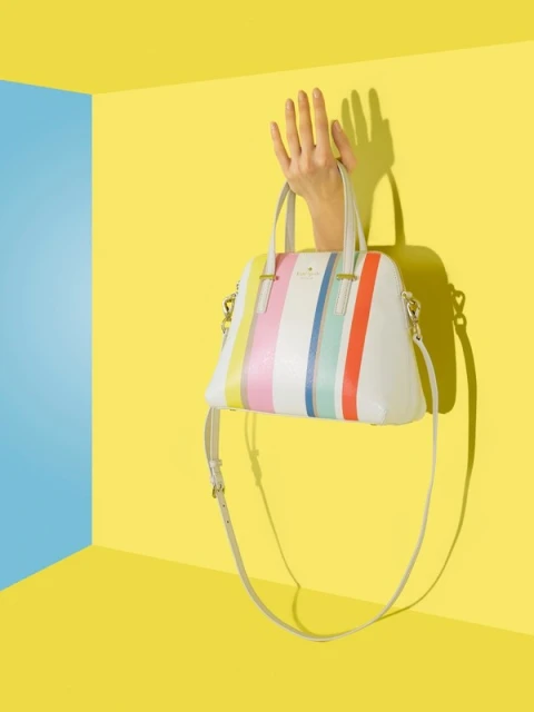 Stylish handbag with colorful stripes held by a hand against a yellow wall with blue corner, creating a vibrant and eye-catching fashion still life.