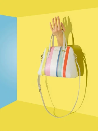 Stylish handbag with colorful stripes held by a hand against a yellow wall with blue corner, creating a vibrant and eye-catching fashion still life.
