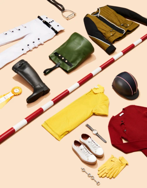 Equestrian-themed apparel and riding gear in a flat lay featuring a riding helmet, boots, gloves, pants, and crop top on a beige background.