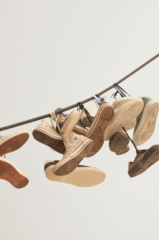 Group of worn canvas sneakers hanging from a rusty metal rod, showcasing worn soles and beige, tan, and brown color palette in studio.