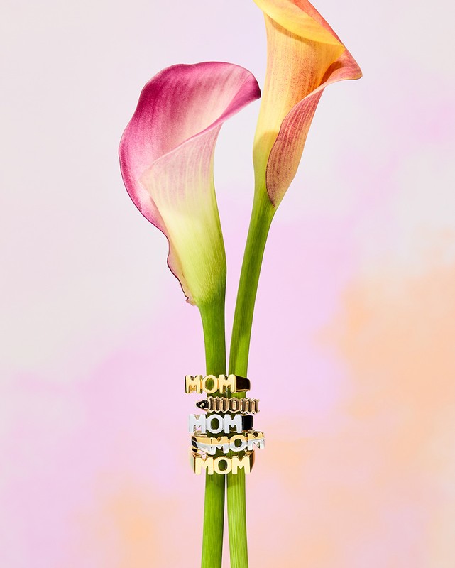 Calla lilies featuring a stack of gold 'MOM' rings, set against a soft-focus pink background, jewelry, and floral arrangement.