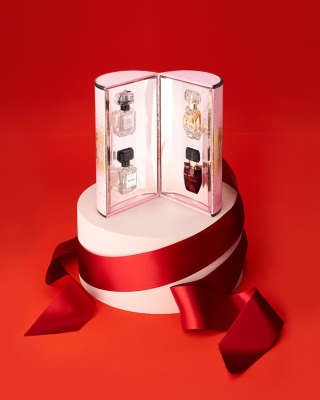 Perfume gift set featuring four miniature perfume bottles in a pink display case, adorned with a red ribbon on a red backdrop.