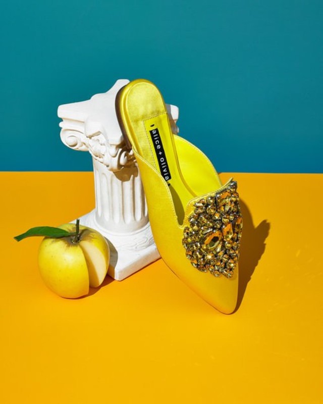 Elegant yellow Alice & Olivia heeled mule with crystal embellishments, resting on a white pillar next to a sliced yellow apple.