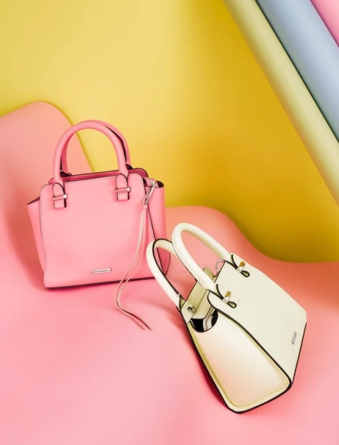 Studio shot of two handbags, one pink and one white, on a pink surface, with yellow and pastel-colored paper rolls in the background.