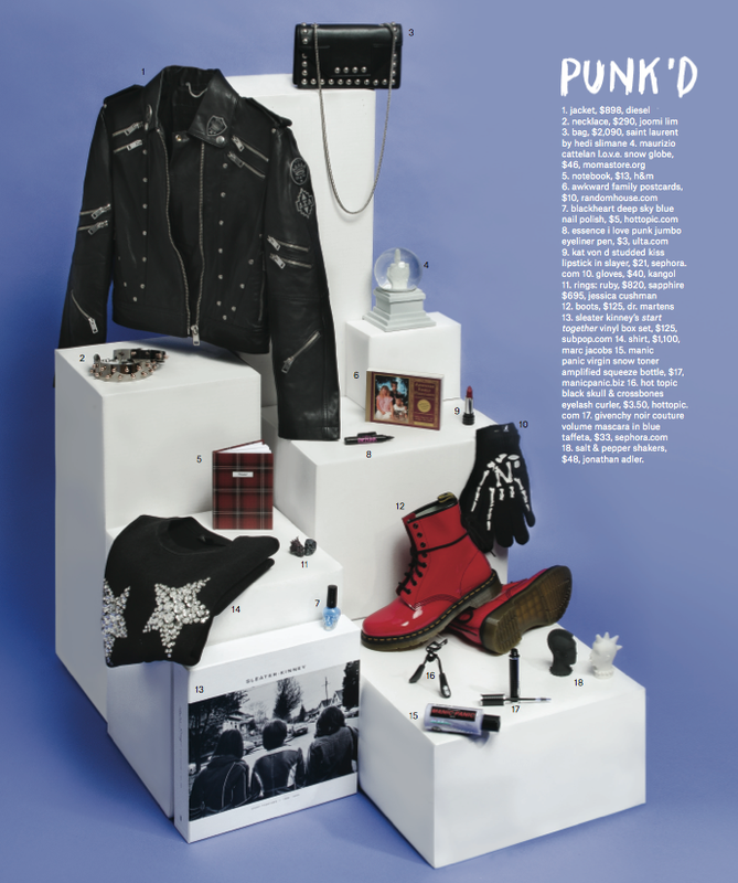 Product still life on a white block display: black leather jacket, black purse, red boots, and various beauty and fashion accessories.