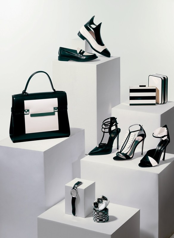 Arrangement of black and white fashion accessories including shoes, a handbag, watches, and a clutch on white geometric display blocks.