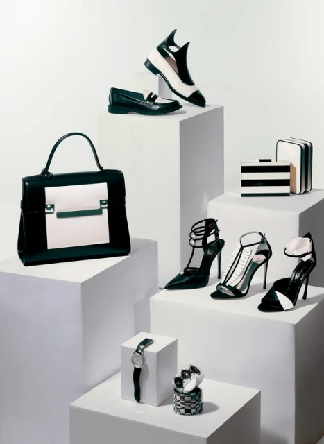 Arrangement of black and white fashion accessories including shoes, a handbag, watches, and a clutch on white geometric display blocks.