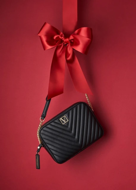 Black chevron-patterned crossbody bag with gold chain strap and 'V' logo, suspended by a red ribbon with a large bow on a red backdrop.