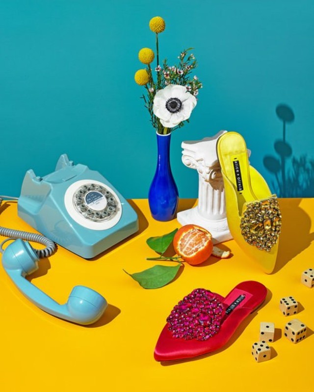 Still life of yellow and red jeweled high heels, telephone, vase with white flowers, citrus fruit, dice set against a teal background.