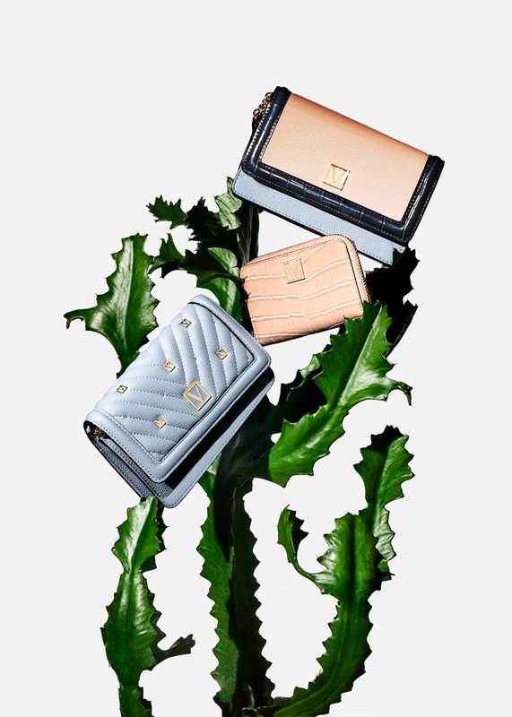 Three pastel-colored clutch purses arranged in a levitating still-life composition with a textured green plant against a white backdrop.