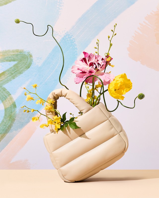 Beige, quilted handbag holding an arrangement of colorful flowers with a painterly pastel background in a studio setting. 