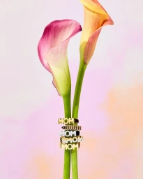 Two tall calla lily stems supporting stacked rings featuring the word mom against a soft pink gradient background