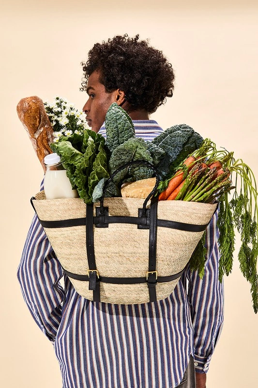 Man wearing striped shirt carrying a straw bag filled with fresh groceries on his back against a pale background