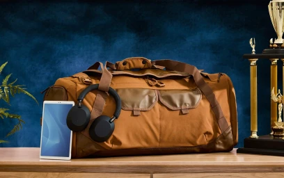 Brown duffel bag resting on a wooden surface with black headphones, a tablet, and a gold trophy.
