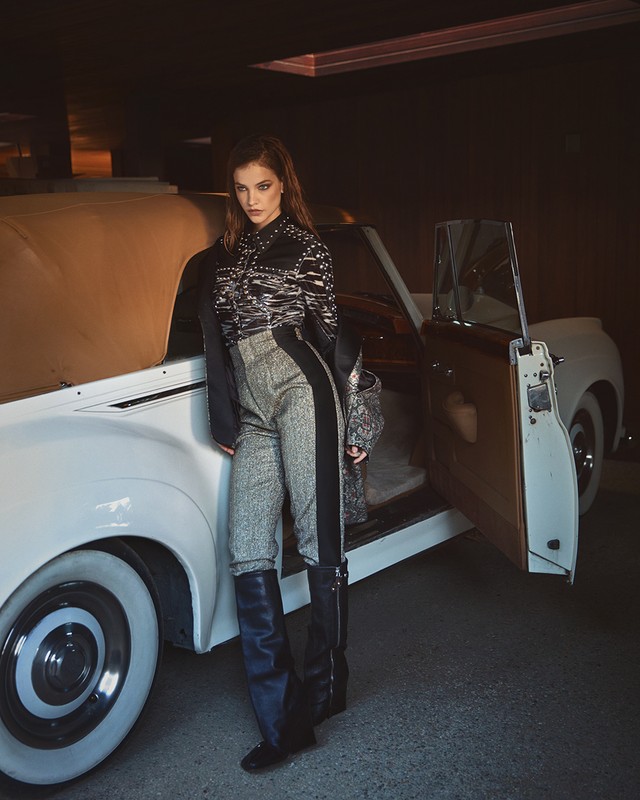 A poised woman in patterned top and high-waisted pants stands beside a vintage car, illuminated by soft light in a dark setting.