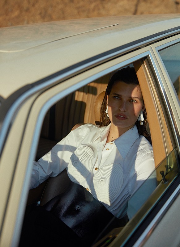 Adult woman poses inside vintage car, wearing white shirt and earrings, bathed in sunlight on a neutral background.