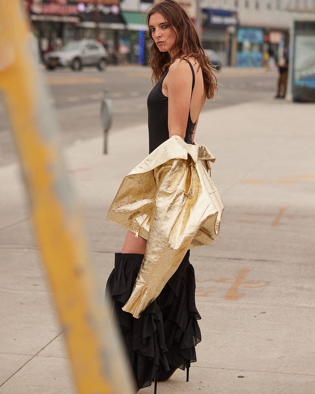Outdoor fashion shoot featuring a woman in a black leotard and skirt, metallic jacket. Urban setting with cars and buildings.