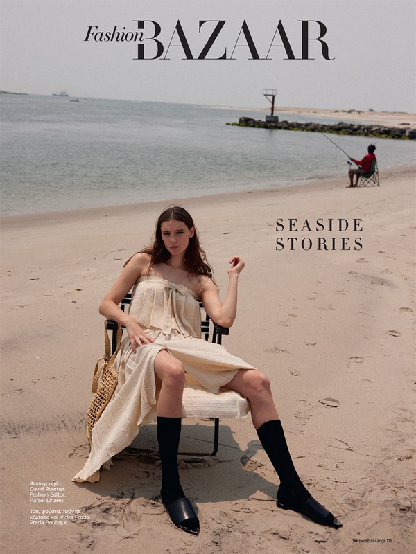 Fashion editorial image features a slender woman in a neutral dress sitting on a beach chair with a fisherman in the distance.