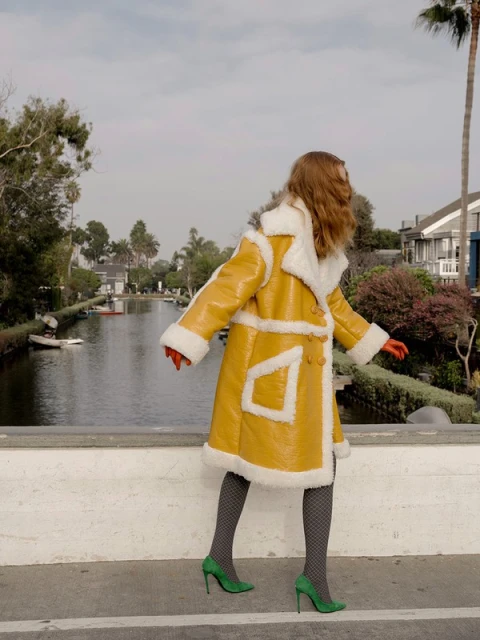 Woman in yellow coat with white fur trim, red gloves, and green heels stands on a bridge over a canal with palm trees.