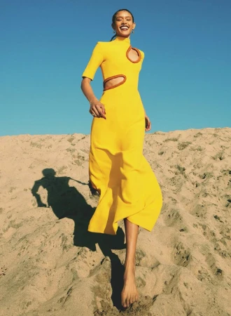 Smiling woman in a yellow cut-out dress walking down a sandy dune under bright sunlight