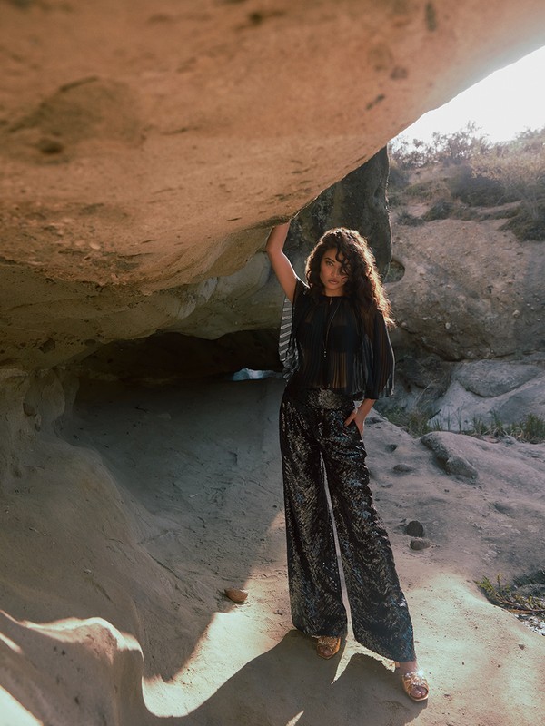 Adult woman with curly hair wearing black top and sequined pants posing in rock cave with warm, even lighting and natural background.